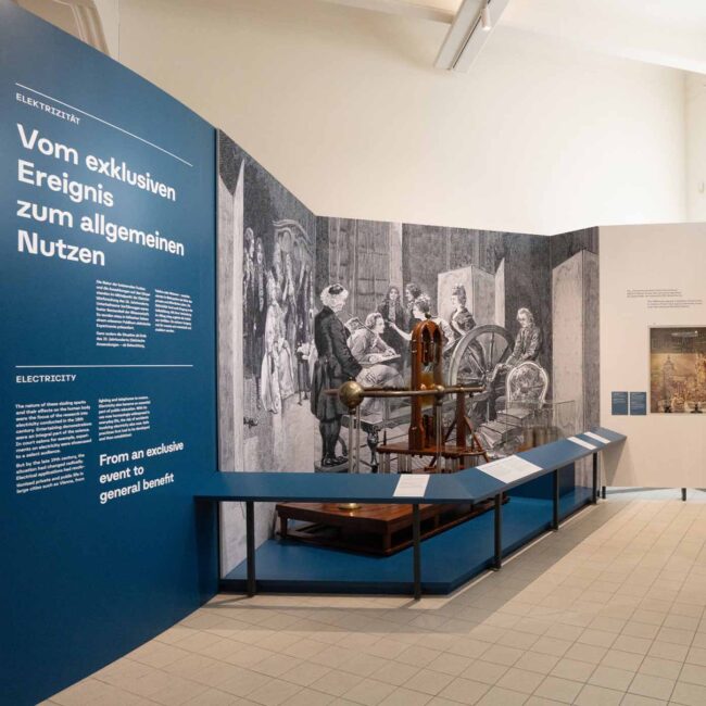 Gesamtansicht der Ausstellungsgrafik „Wirtschaft im Wandel" im Technischen Museum Wien – Blauer Themenbereich Elektrizität mit bedruckten Stoffbespannungen in Alu-Frames, Folienplots und Beschriftungen, umgesetzt durch print-art 2026
